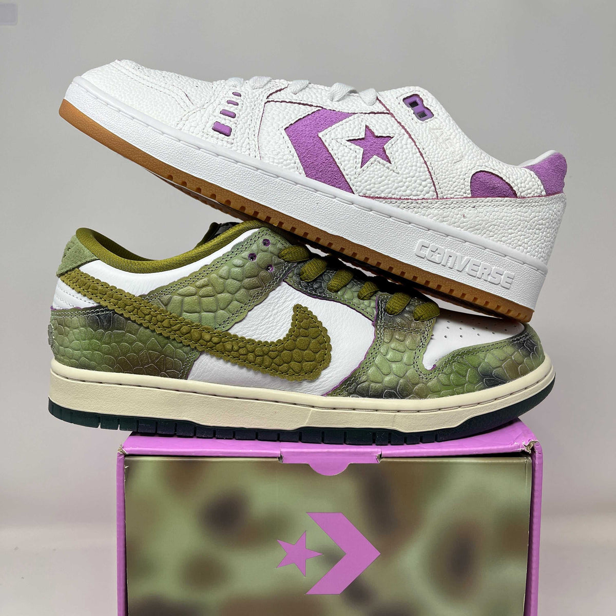 Nike SB Dunk Low sneakers with green reptile-textured panels, cream leather, olive swoosh and dark green soles, paired with a white and purple Converse shoe.